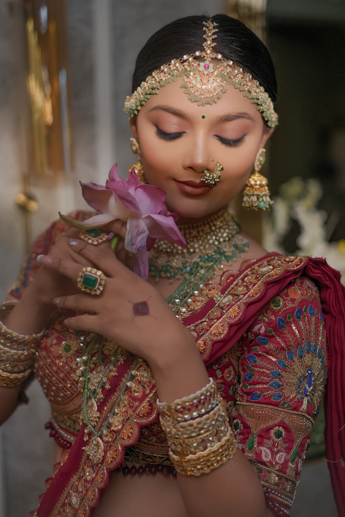 Bride in a deep red embroidered lehenga wearing a pearl and emerald maang tikka, kundan nath and layered necklaces, eyes closed, holding a lotus flower